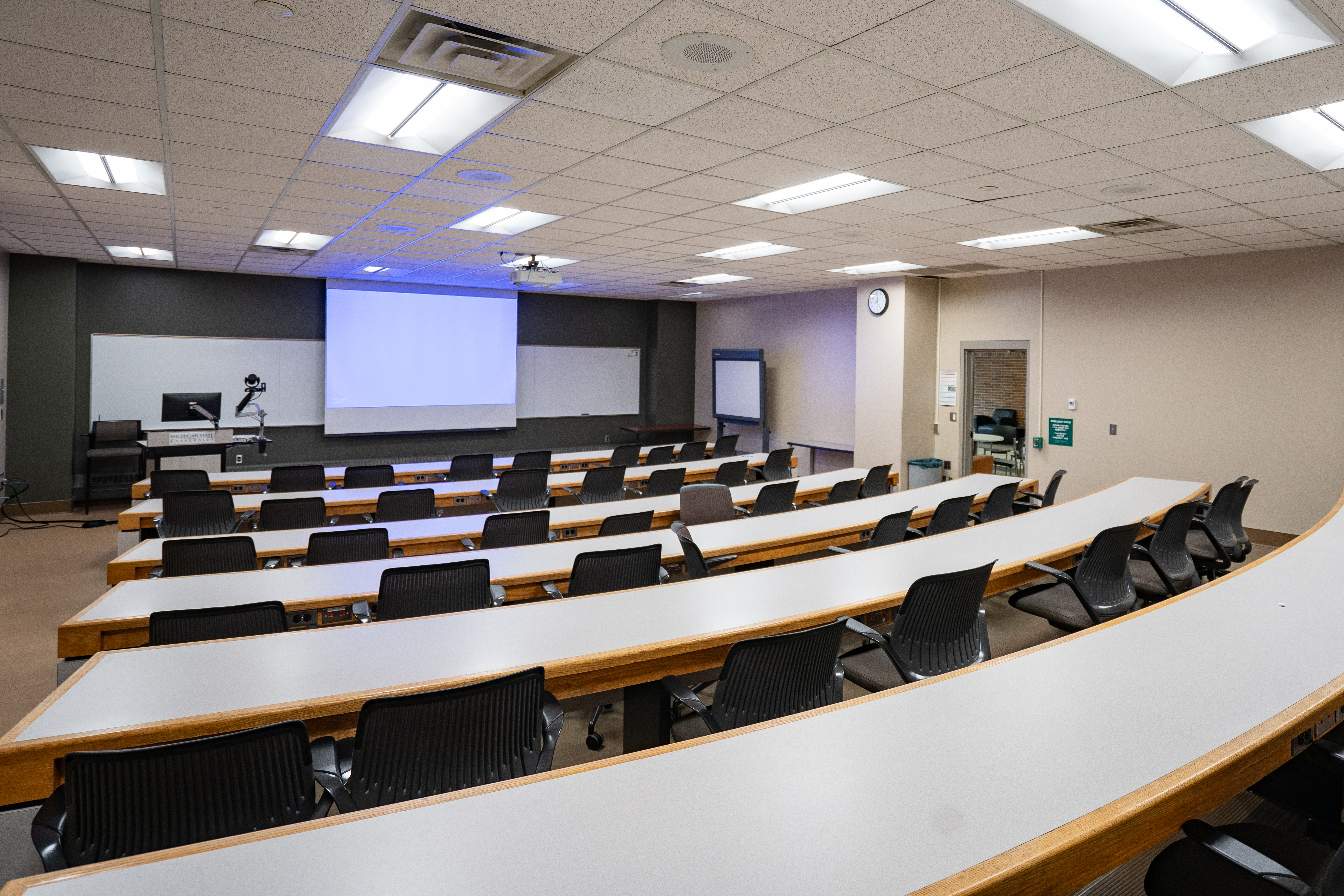 A classroom of 5 table rows with chairs facing a projector screen and podium