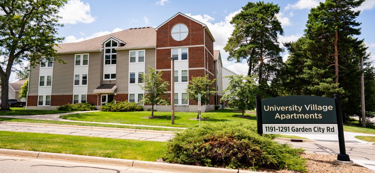 Multi-story apartment building with brick and siding exterior, featuring numerous windows and a central entrance with steps. The building is surrounded by trees and neatly maintained lawns. A sign in the foreground reads 'University Village Apartments 1191-1291 Garden City Rd.' The sky is partly cloudy, and the scene is bright and sunny.