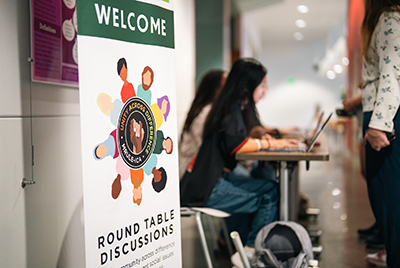 Photo with MRULE banner in foreground and Intercultural Aides sitting at a table and checking people into for a round-table discussions in background