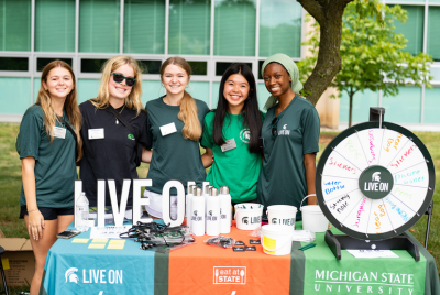 image of five live on campus crew members standing at a table with a prize wheel and Live On merch