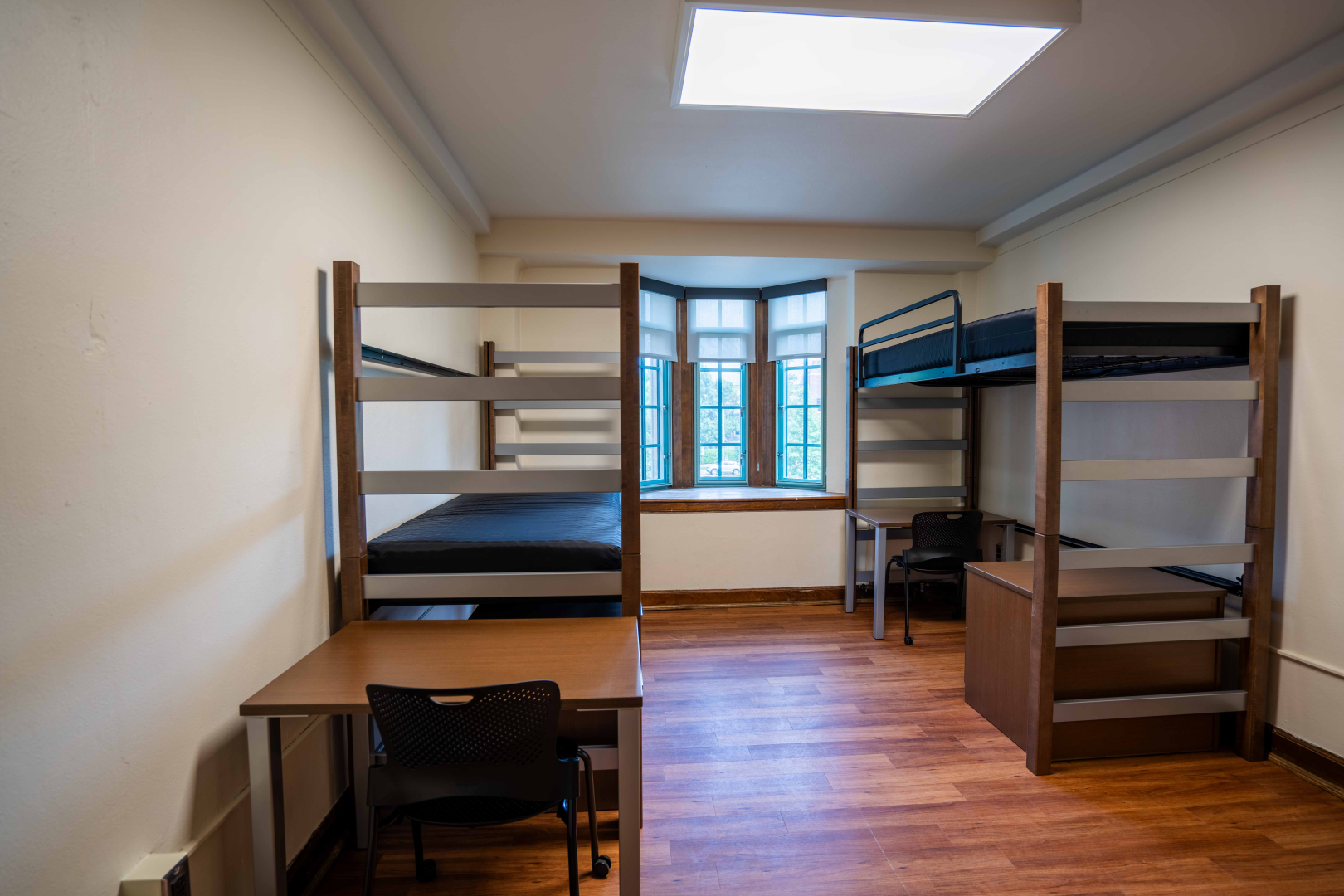 Photo from entrance door facing window of a sample room in Campbell Hall. Shows unfurnished furniture set-up of a lofted and a trundled bed, two desks and chairs, and two small dressers.