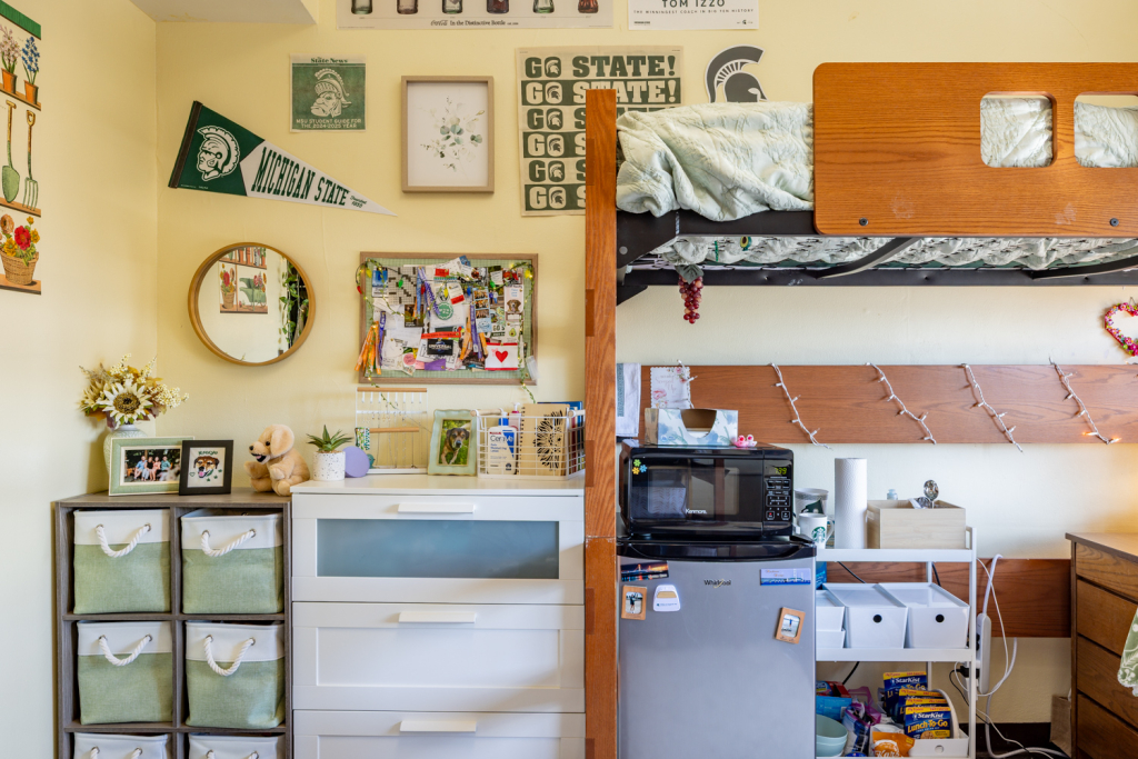 A decorated residence hall room features a lofted wooden bed with light green bedding, Michigan State University-themed wall décor, and a cozy storage area below. A white dresser is topped with plants, framed photos, and storage containers. Nearby are a mini refrigerator with a microwave on top, open shelving stocked with snacks and supplies, string lights, and a bulletin board filled with photos, notes, and mementos. A round wall mirror and green-and-white storage bins add to the organized, personalized space.