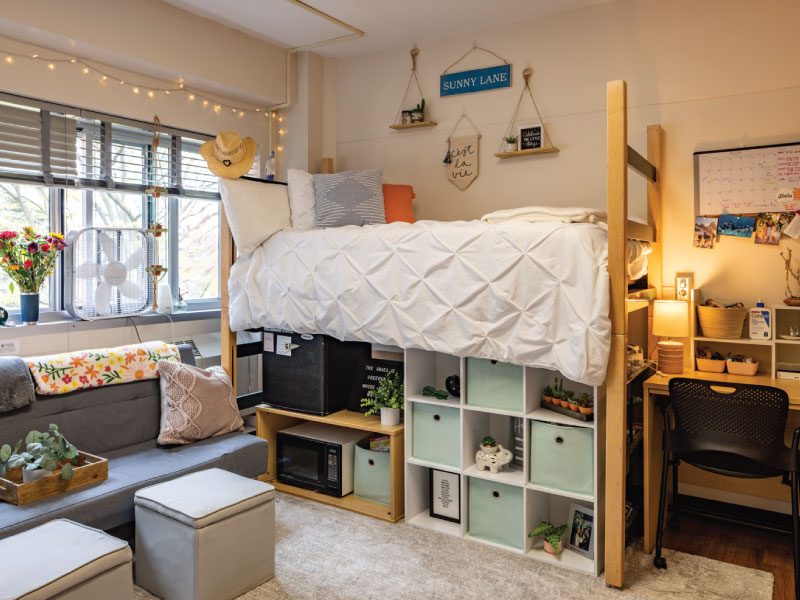 Cozy dorm room with a large window, comfy chair, microwave, fridge, desk, and decorated walls.