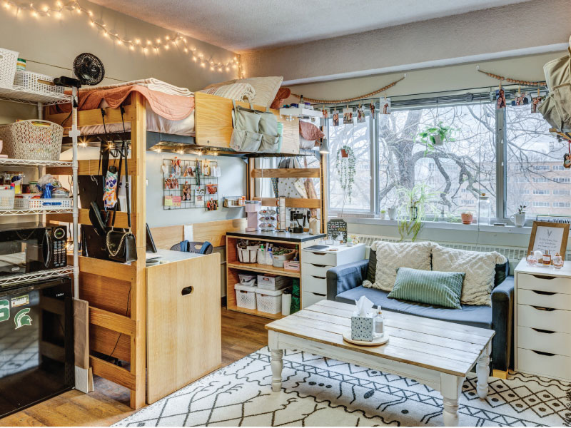 Cozy dorm room with a large window, comfy chair, microwave, fridge, desk, and decorated walls.