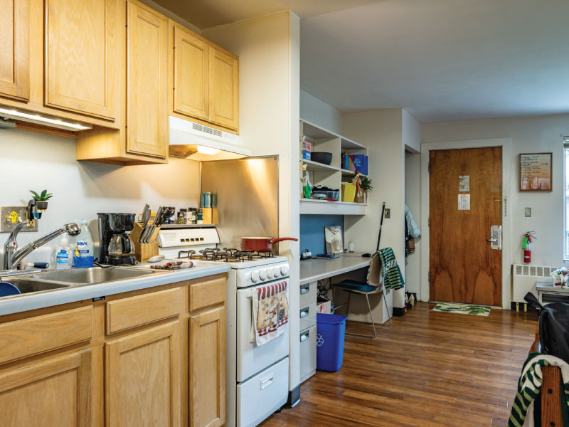 A small kitchen and study area with wooden cabinets, a gas stove, built-in shelving, a desk, and an entrance door.